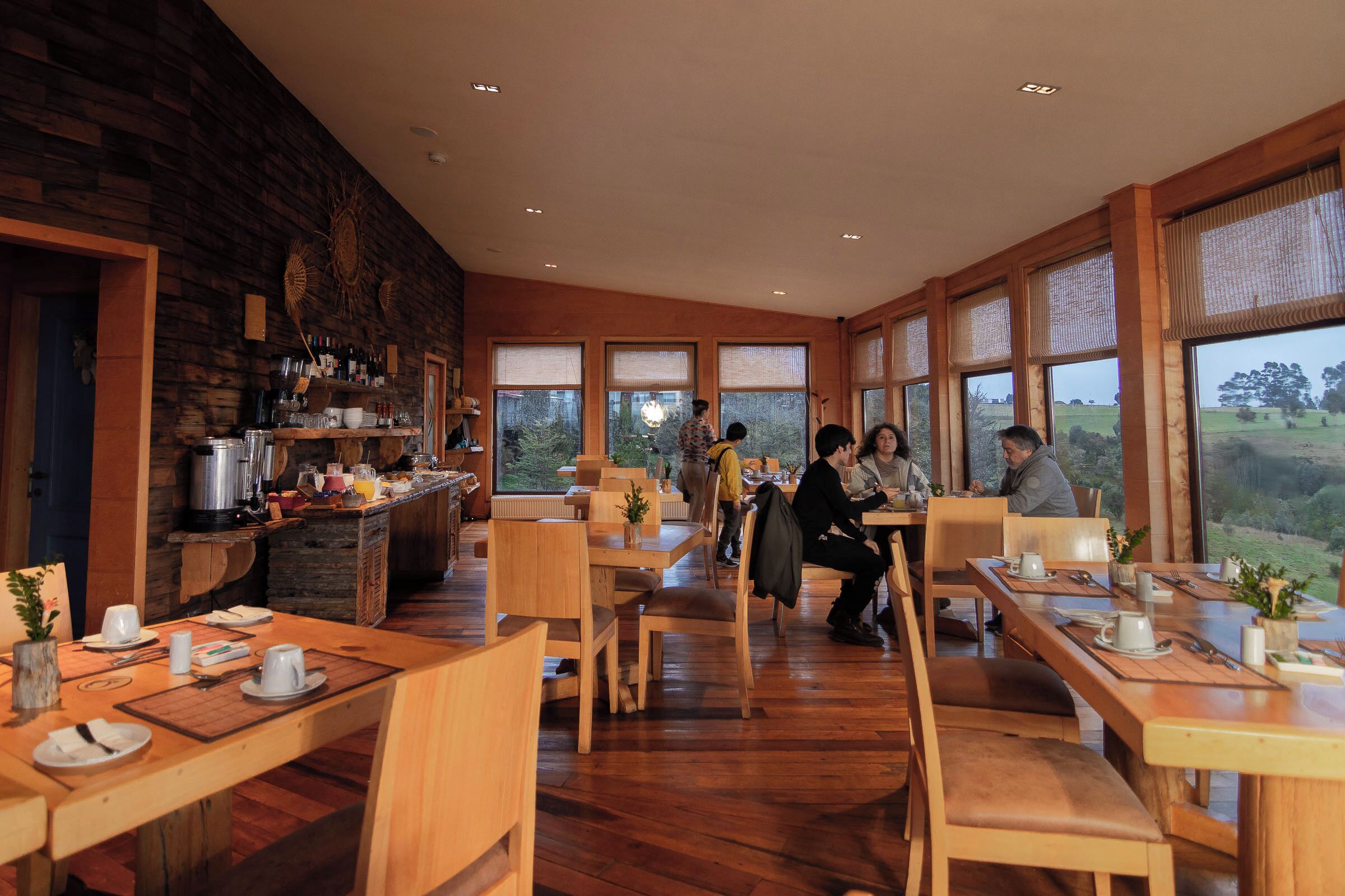A cozy restaurant with wooden furniture and large windows offering a view of the outdoors.