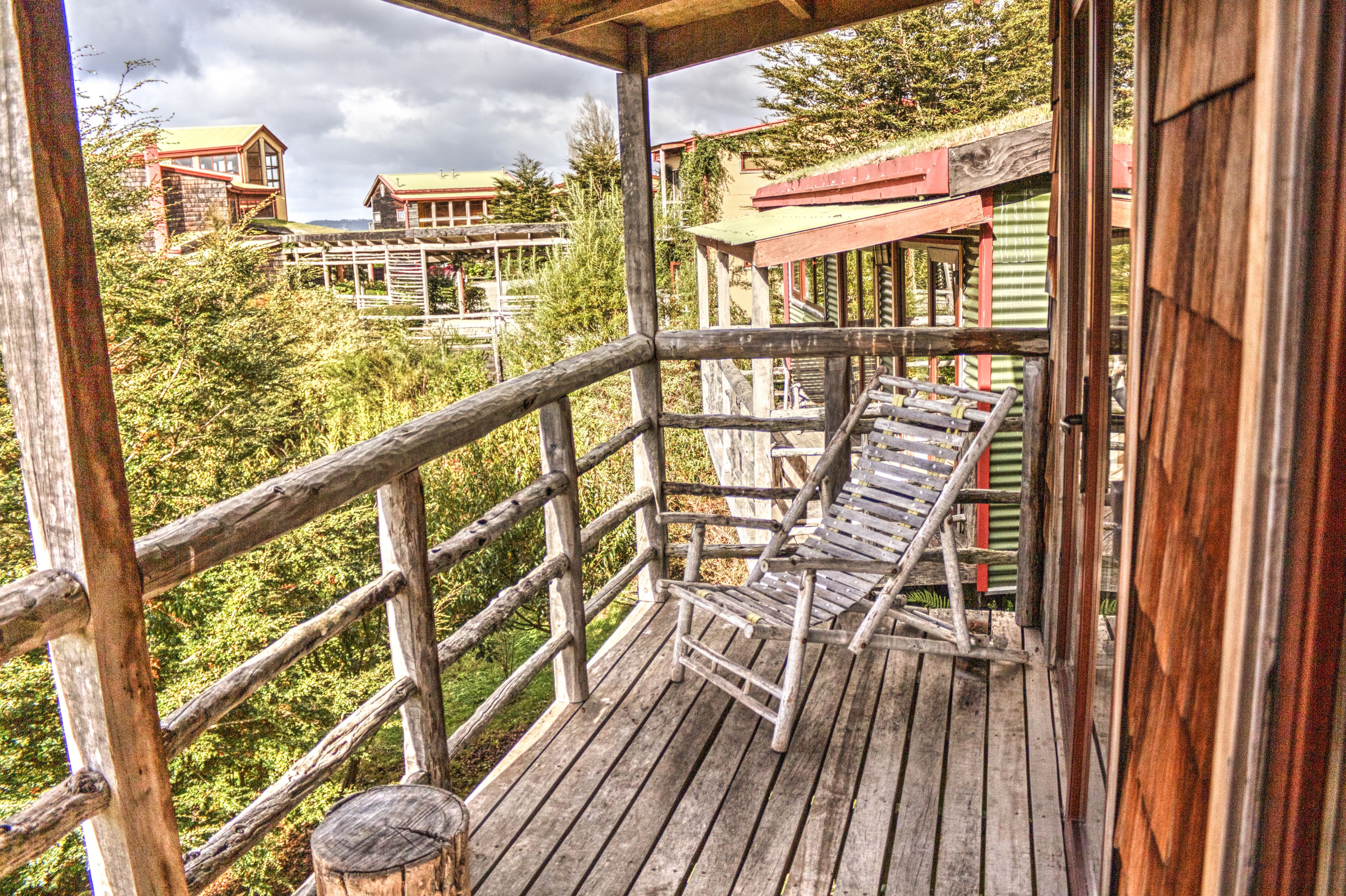 A wooden deck with a bench, surrounded by a wooden railing, overlooking a forested area with buildings in the background.