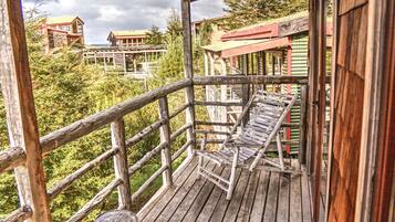 A wooden deck with a bench, surrounded by a wooden railing, overlooking a forested area with buildings in the background.