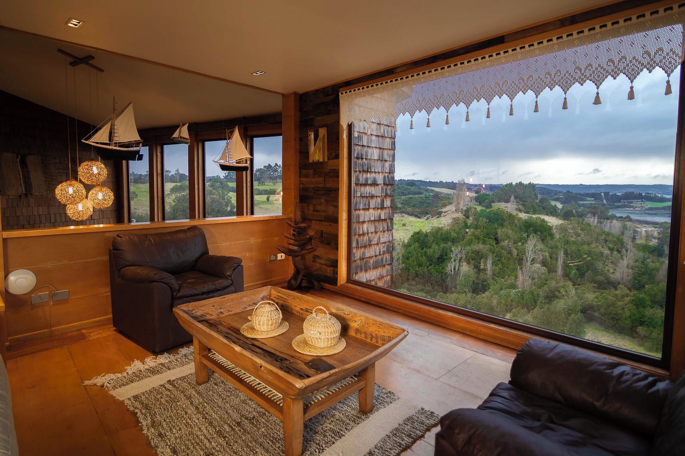 A spacious living room with a large window offering a view of a forested landscape. A leather sofa and a wooden coffee table with decorative items are positioned in front of the window.