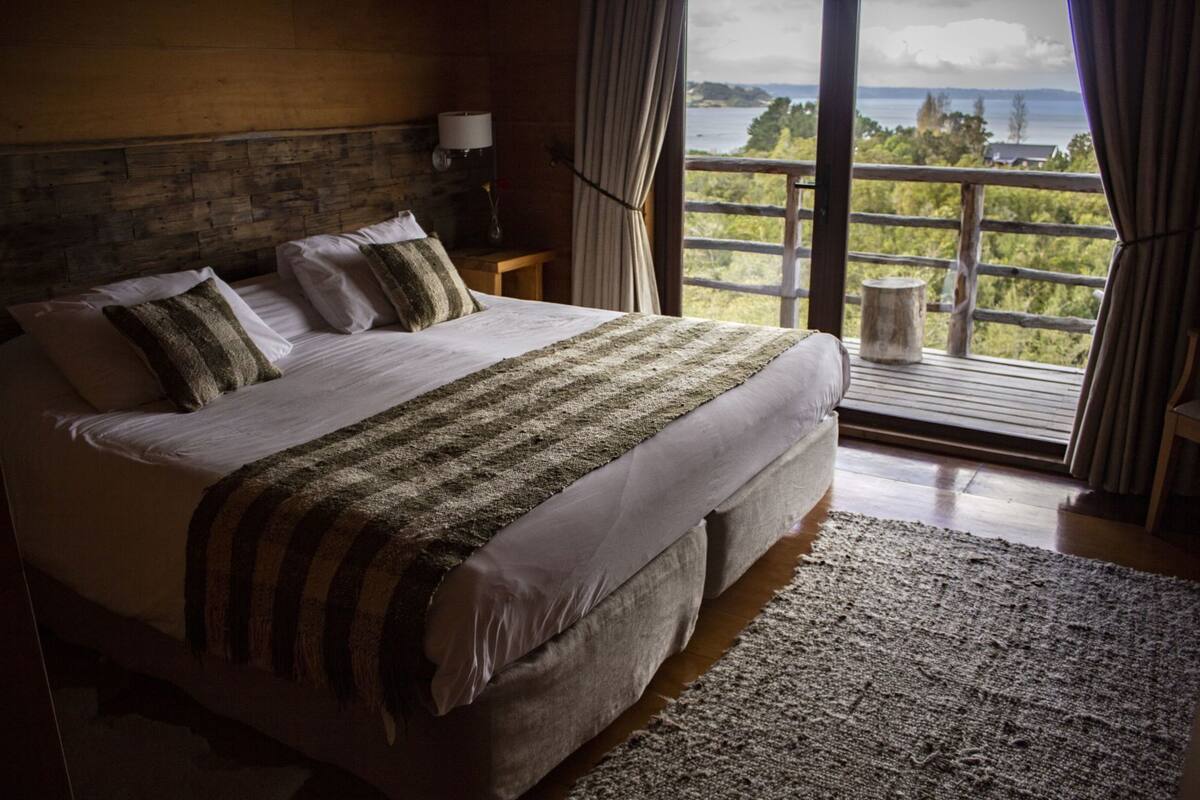 A large bed with a view of the outdoors, a wooden headboard, and a small table with a lamp.