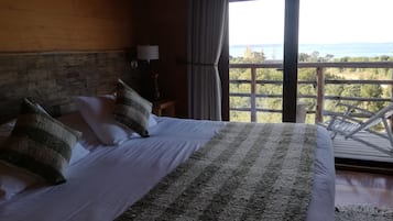 A bedroom with a large bed, a wooden headboard, a nightstand with a lamp, and a view of the outdoors through a large window.