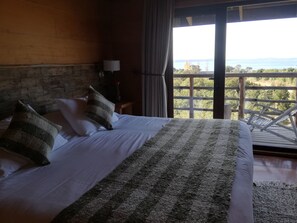 A bedroom with a large bed, a wooden headboard, a nightstand with a lamp, and a view of the outdoors through a large window.