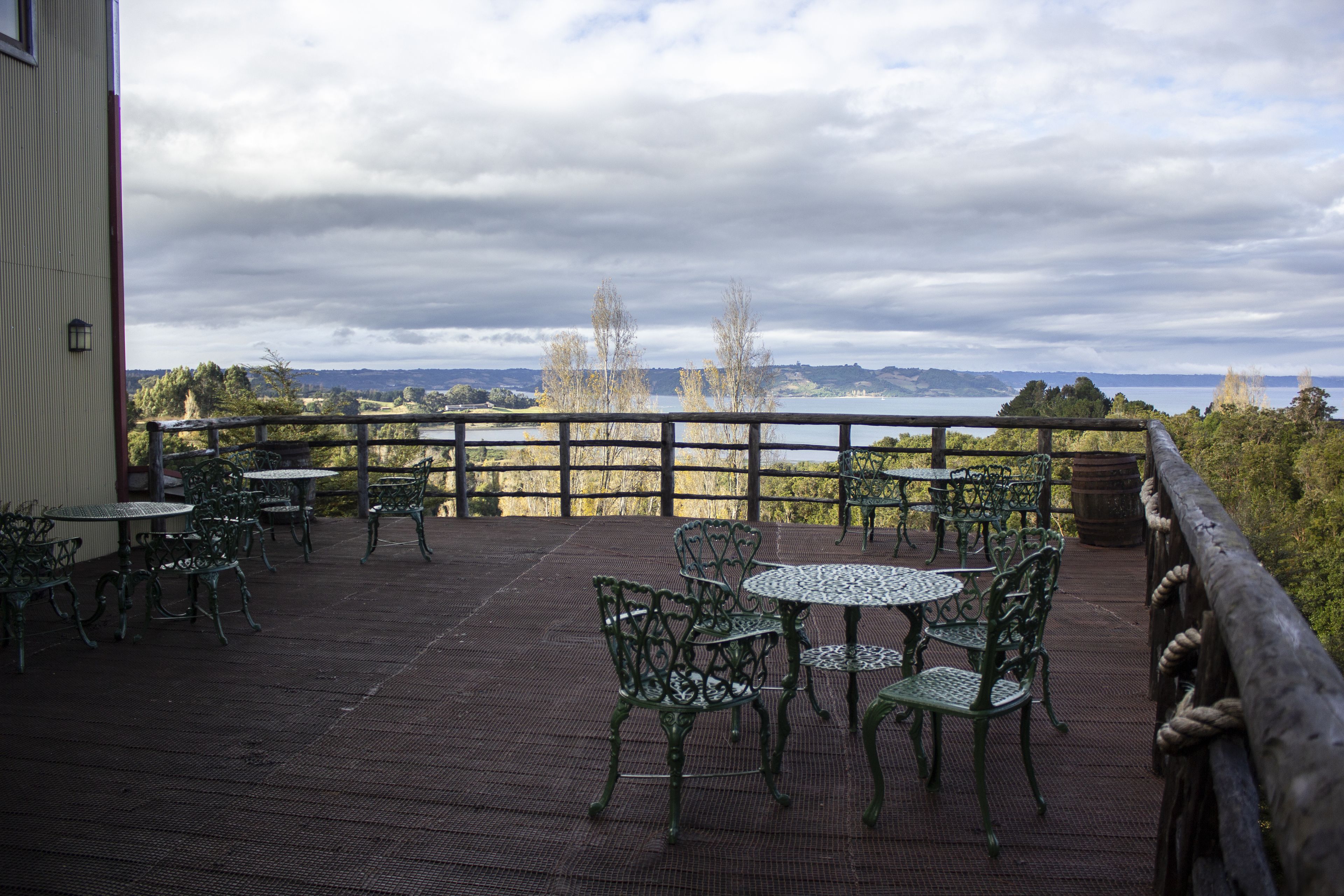 Un espacio al aire libre techado con mesas y sillas de metal, baranda de madera y vista a un lago y colinas al fondo.
