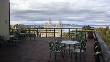 Un espacio al aire libre techado con mesas y sillas de metal, baranda de madera y vista a un lago y colinas al fondo.