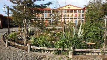 Un hotel de varios pisos, tejado rojo y jardín con plantas diversas y cerca de madera.