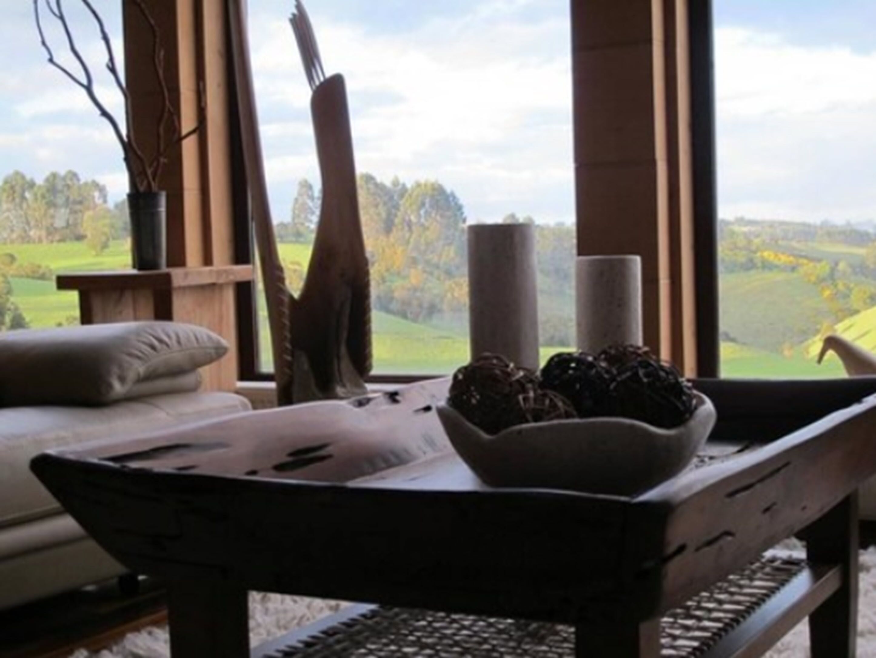A room with a large window offering a view of a green landscape. There is a wooden table with a bowl of fruit and a candle.