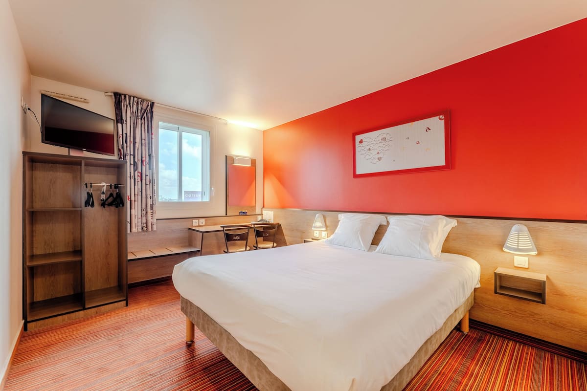 A hotel room with a bed, desk, chair, wardrobe, and a TV.