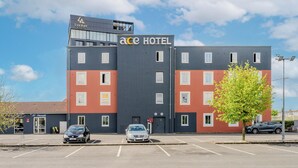 Un hotel con un diseño moderno, que destaca un cartel grande que dice 'ACE HOTEL'. Hay varios espacios de estacionamiento al frente, y se ve un árbol a la derecha.