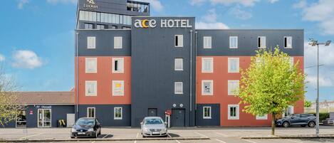 A hotel with a modern design, featuring a prominent 'ACE HOTEL' sign. There are multiple parking spaces in front, and a tree is visible to the right.