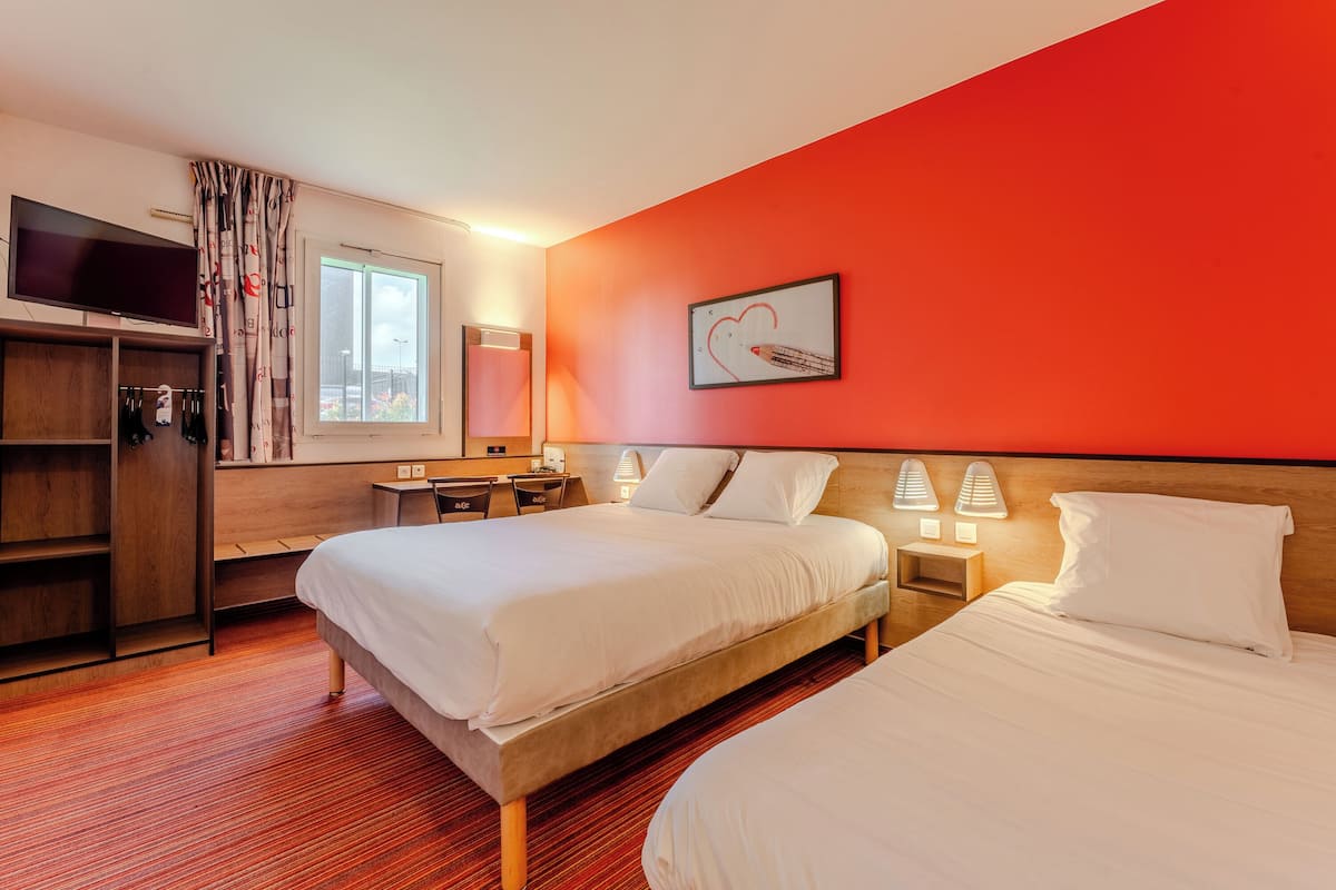 A hotel room with two beds, a TV, a desk with a chair, a window, and a red accent wall.