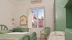 A room with two beds, a desk, and a chair. There is a window with a view of buildings outside. - Hotel Del Postiglione (Ischia)