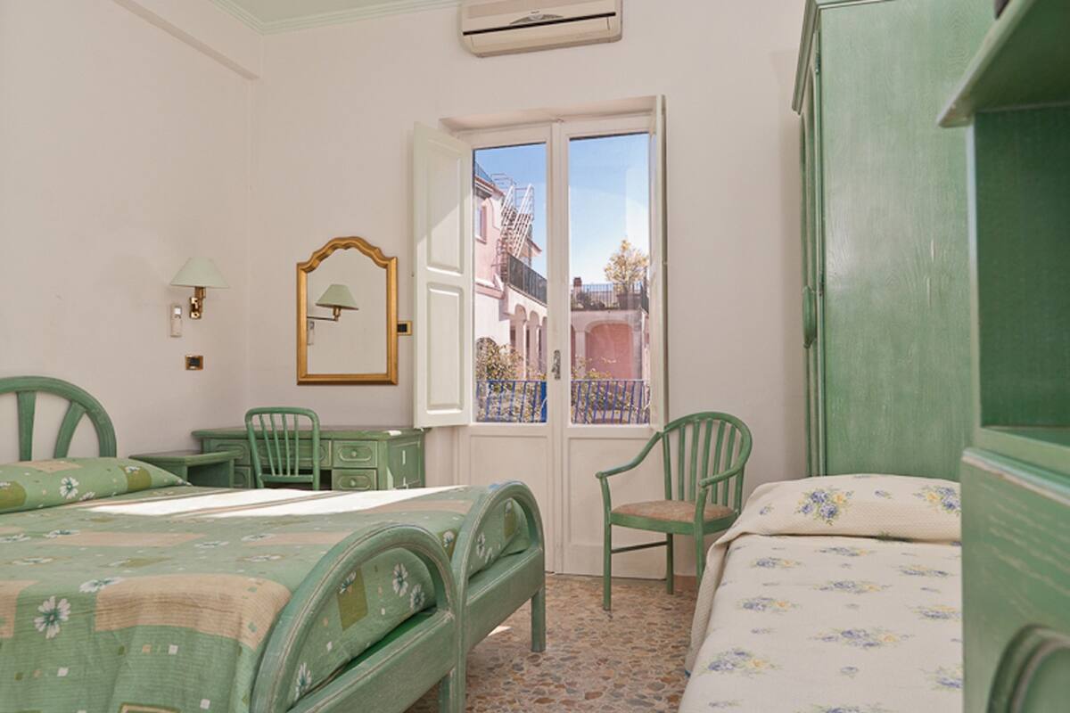 A room with two beds, a desk, and a chair. There is a window with a view of buildings outside.