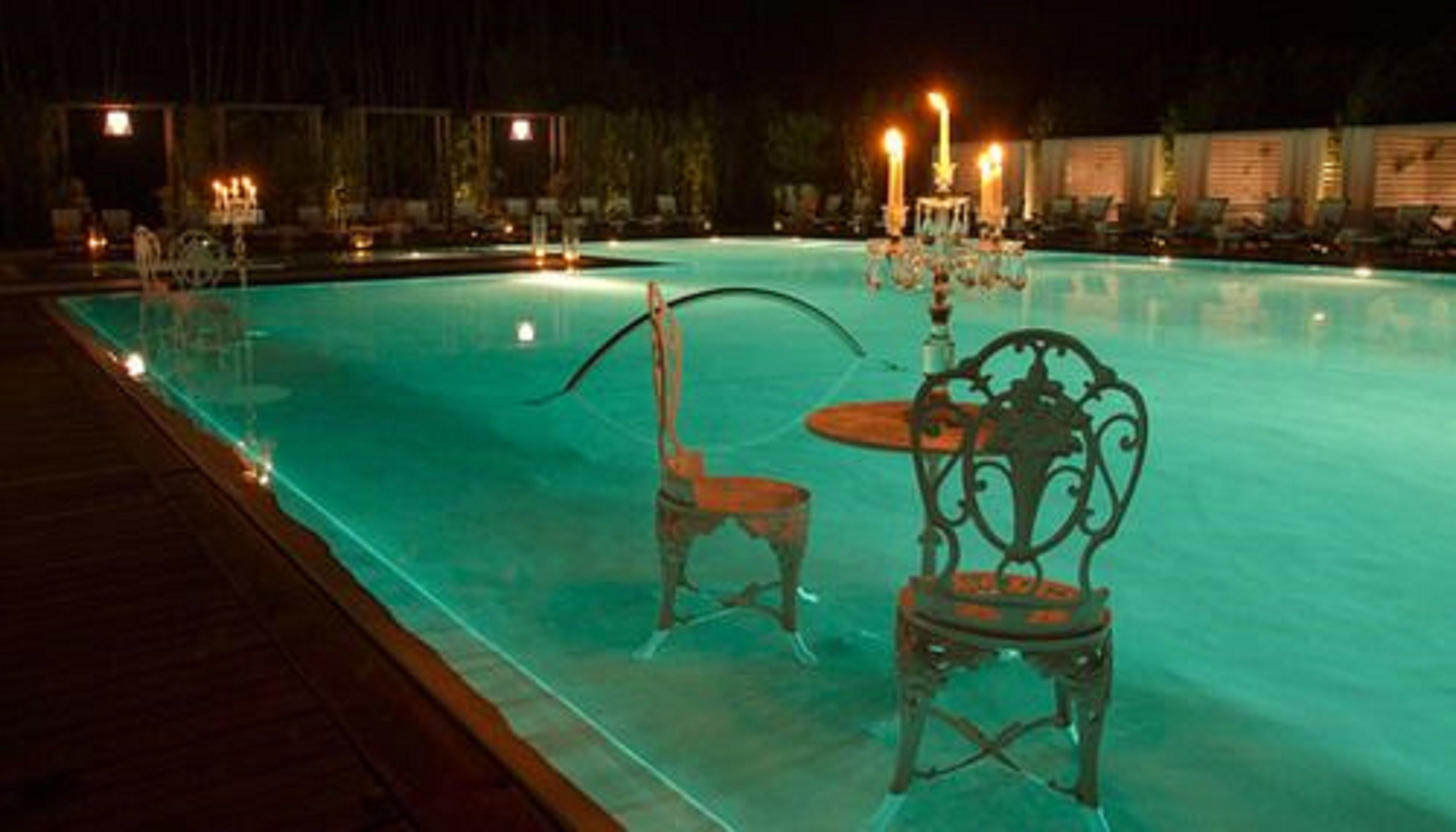 Vista nocturna de una piscina con dos sillas y una mesa, rodeada de sillones y velas.