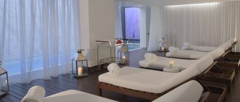 Un cuarto de spa con camastros, una mesa de vidrio con velas y vistas a una zona de piscina.