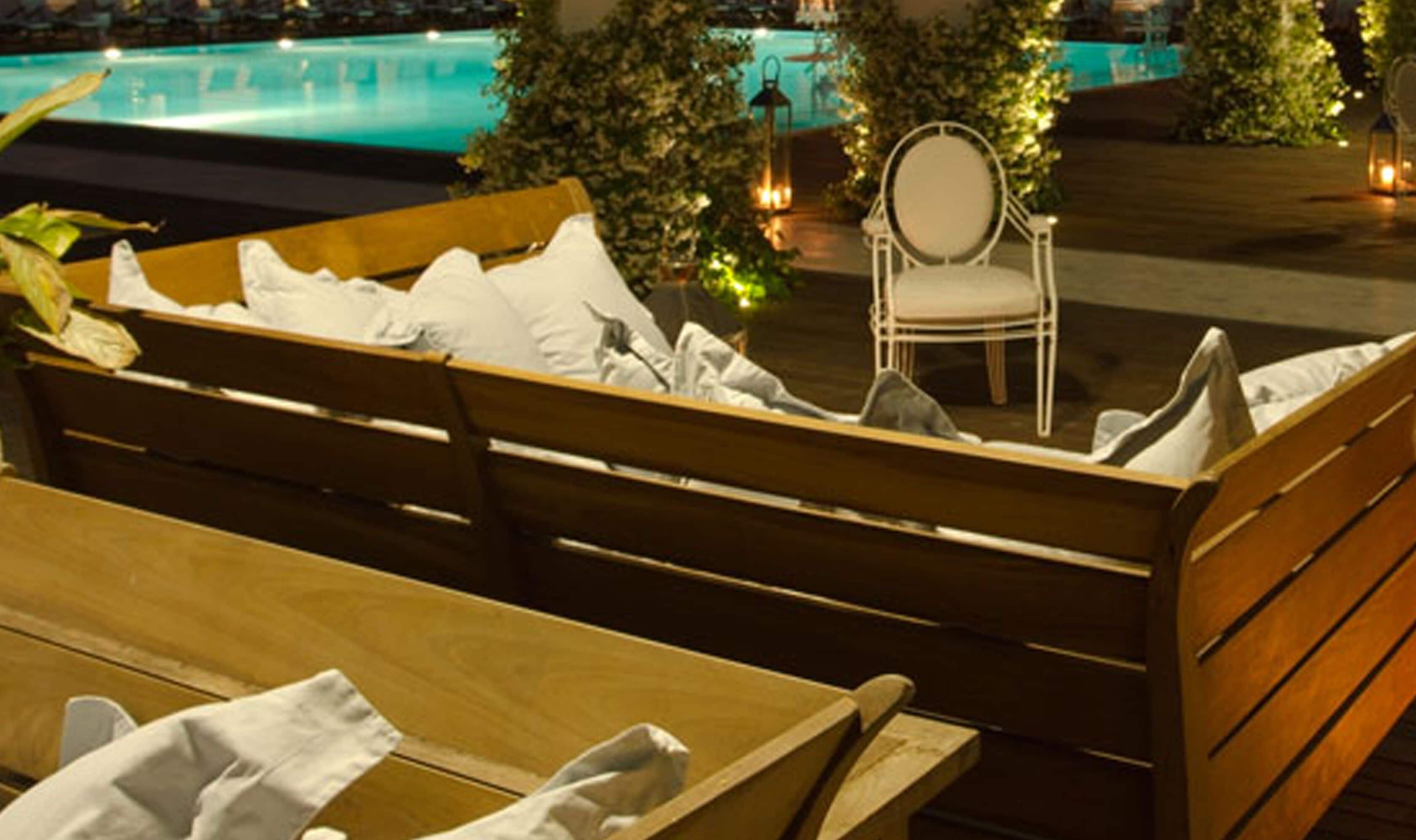 Un banc en bois avec des coussins et une chaise, placés au bord d’une piscine, la nuit.