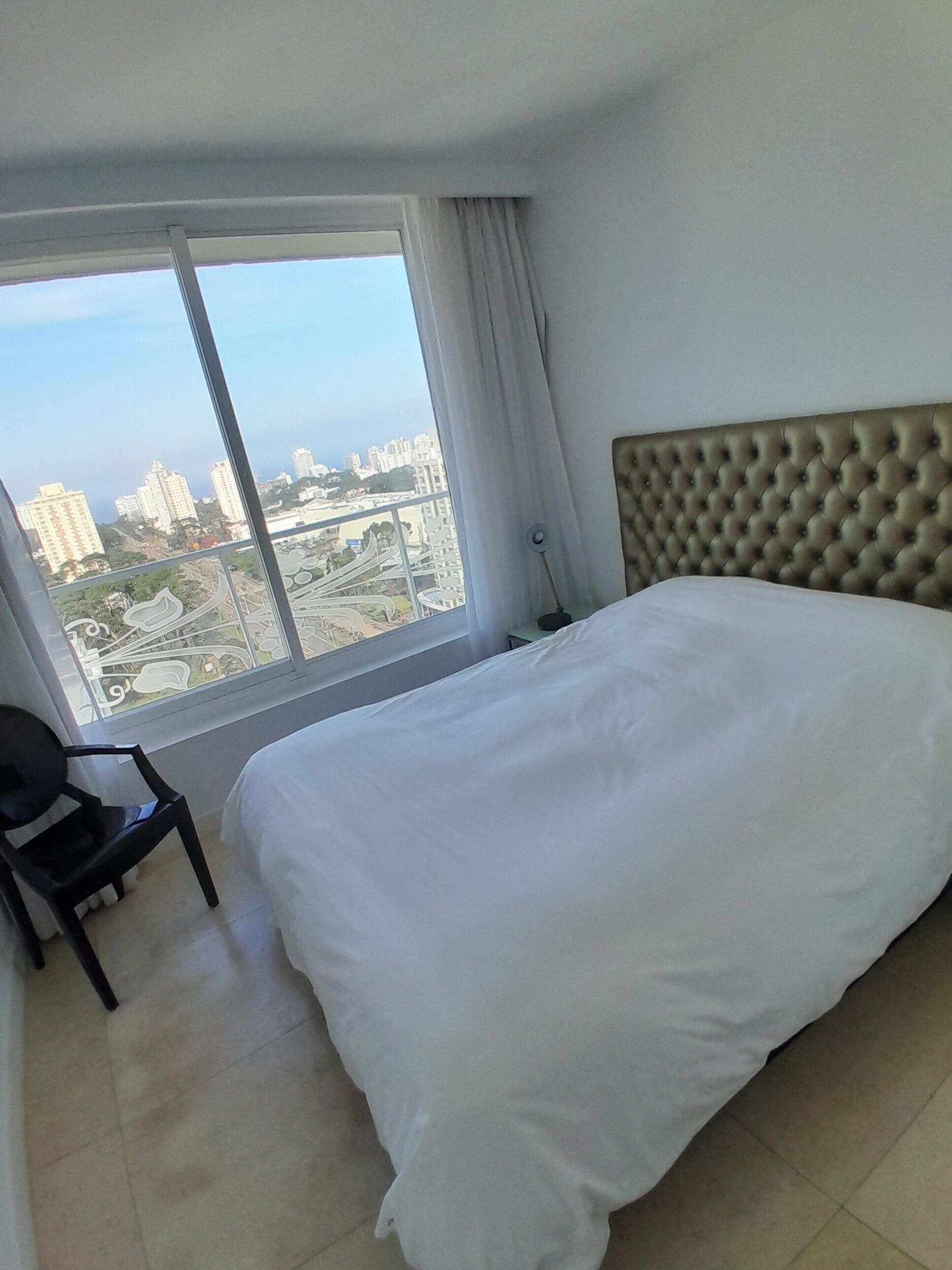 A hotel room with a large bed, a chair, a nightstand, and a window offering a city view.