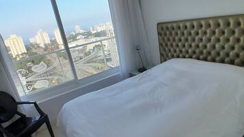 A hotel room with a large bed, a chair, a nightstand, and a window offering a city view.