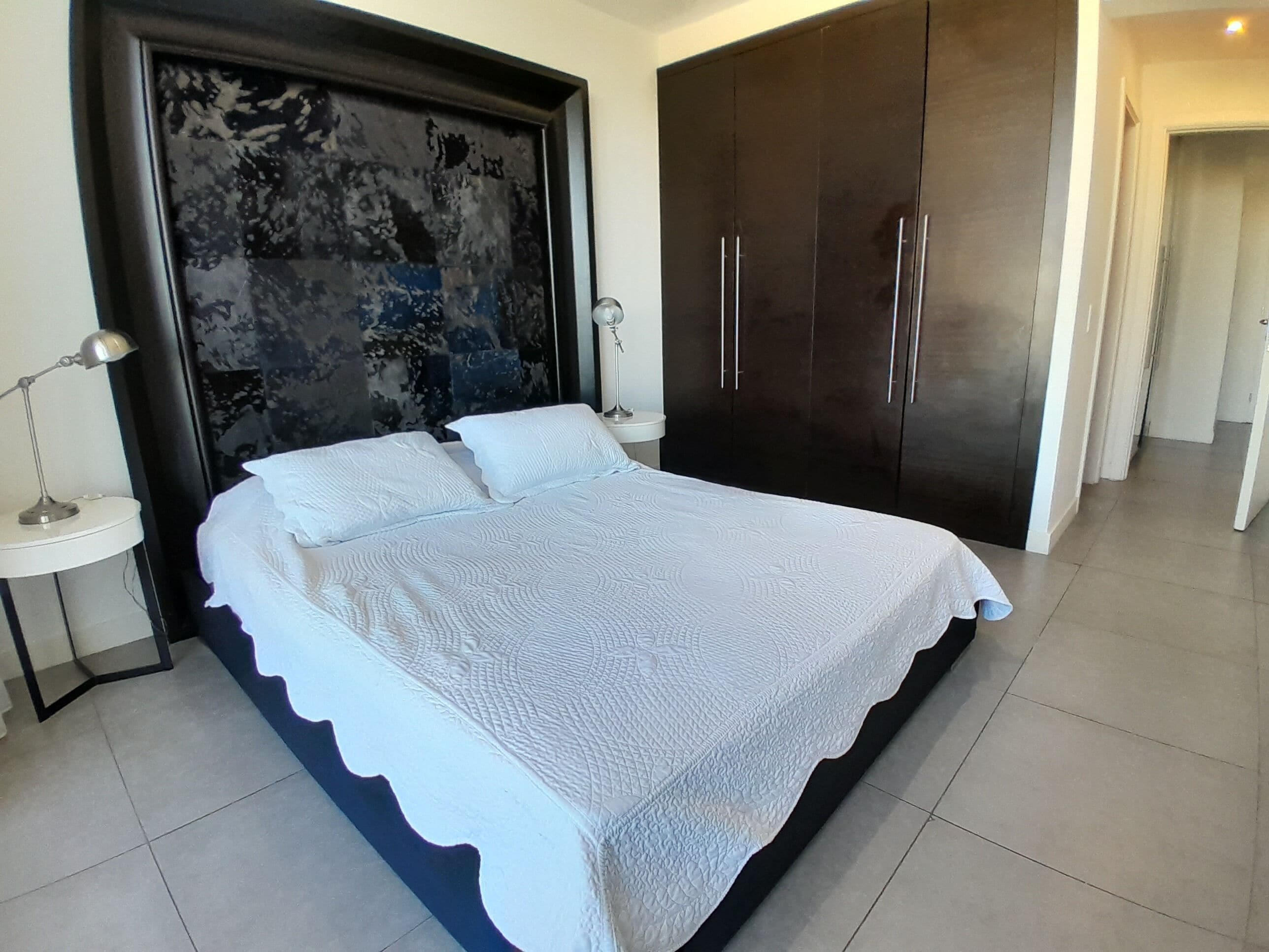 A bedroom with a large bed, two bedside tables, a dark wardrobe, and a decorative headboard.