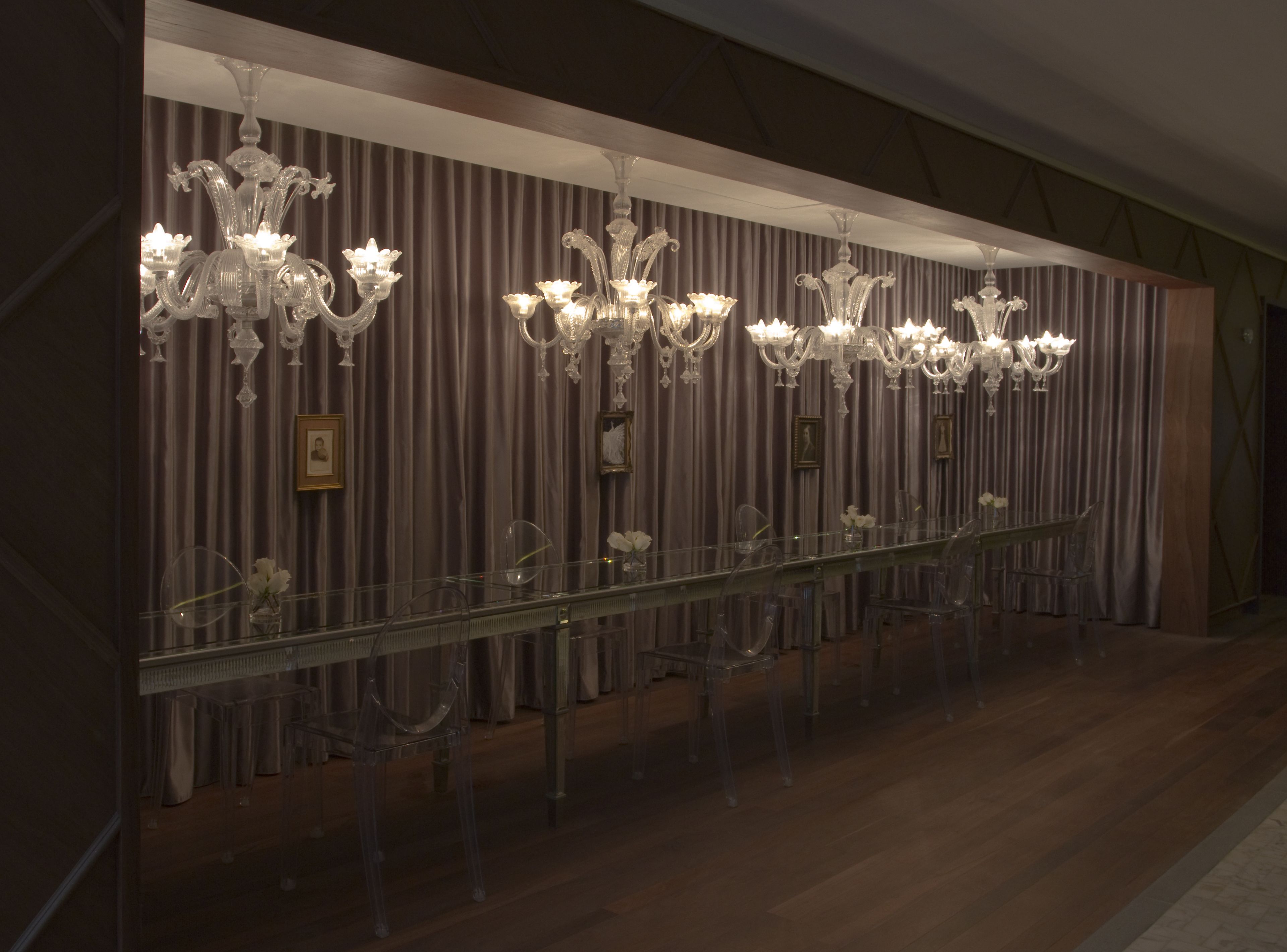 A long, clear glass table with chairs, flanked by ornate chandeliers and a draped curtain backdrop.