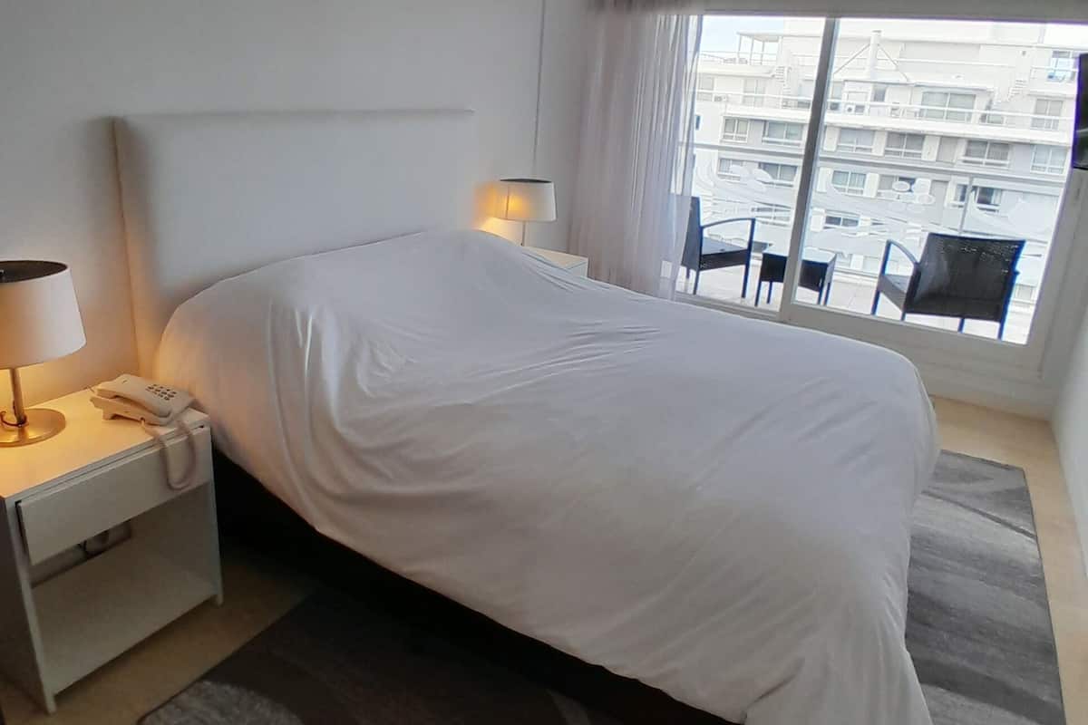 Hotellihuone, jossa on suuri sänky, yöpöytä lampun kanssa ja ikkunasta avautuva näkymä kaupunkiin.
