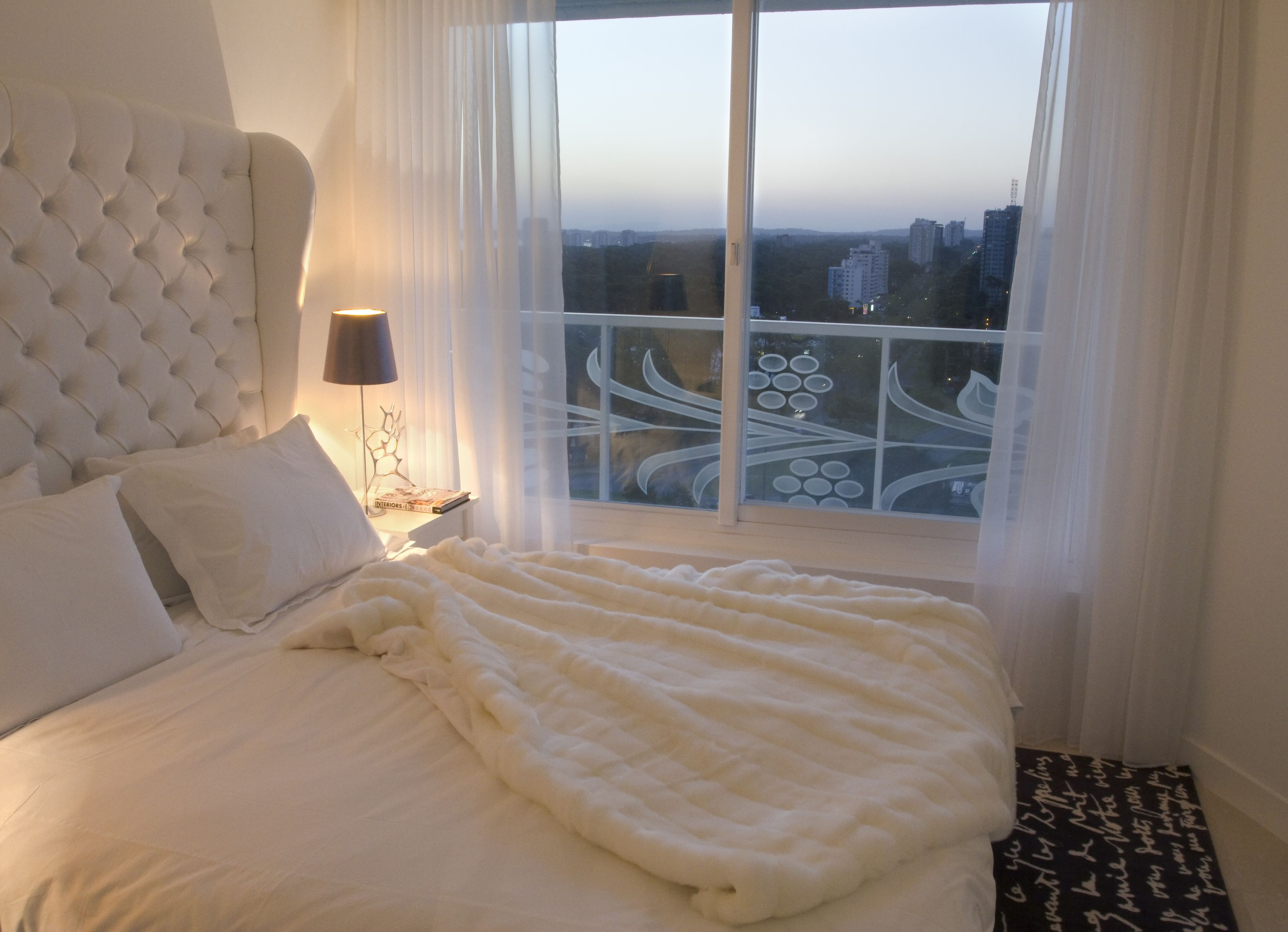 Un dormitorio con una cama grande, un cabecero, una mesita de noche con una lámpara y vistas a un paisaje urbano desde un balcón.