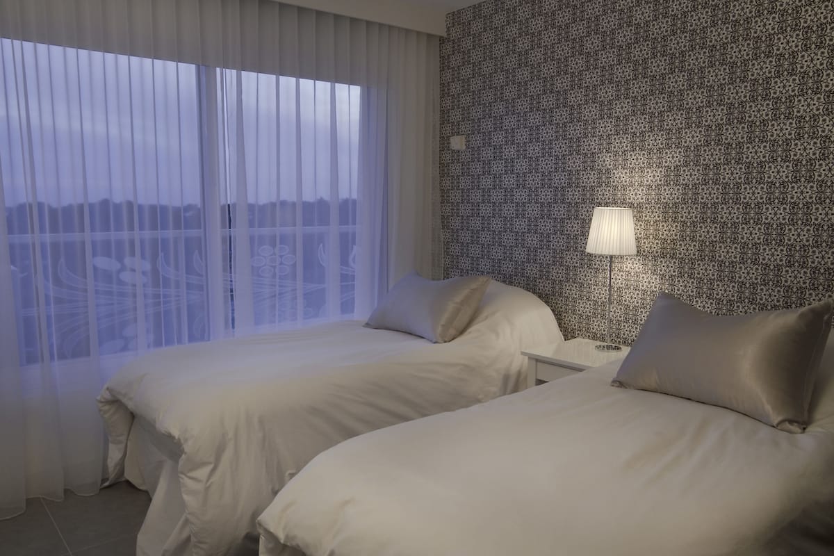 Hotellihuone, jossa on kaksi sänkyä, suuri ikkuna läpikuultavilla verhoilla, kuvioitu seinä ja yöpöytälamppu.