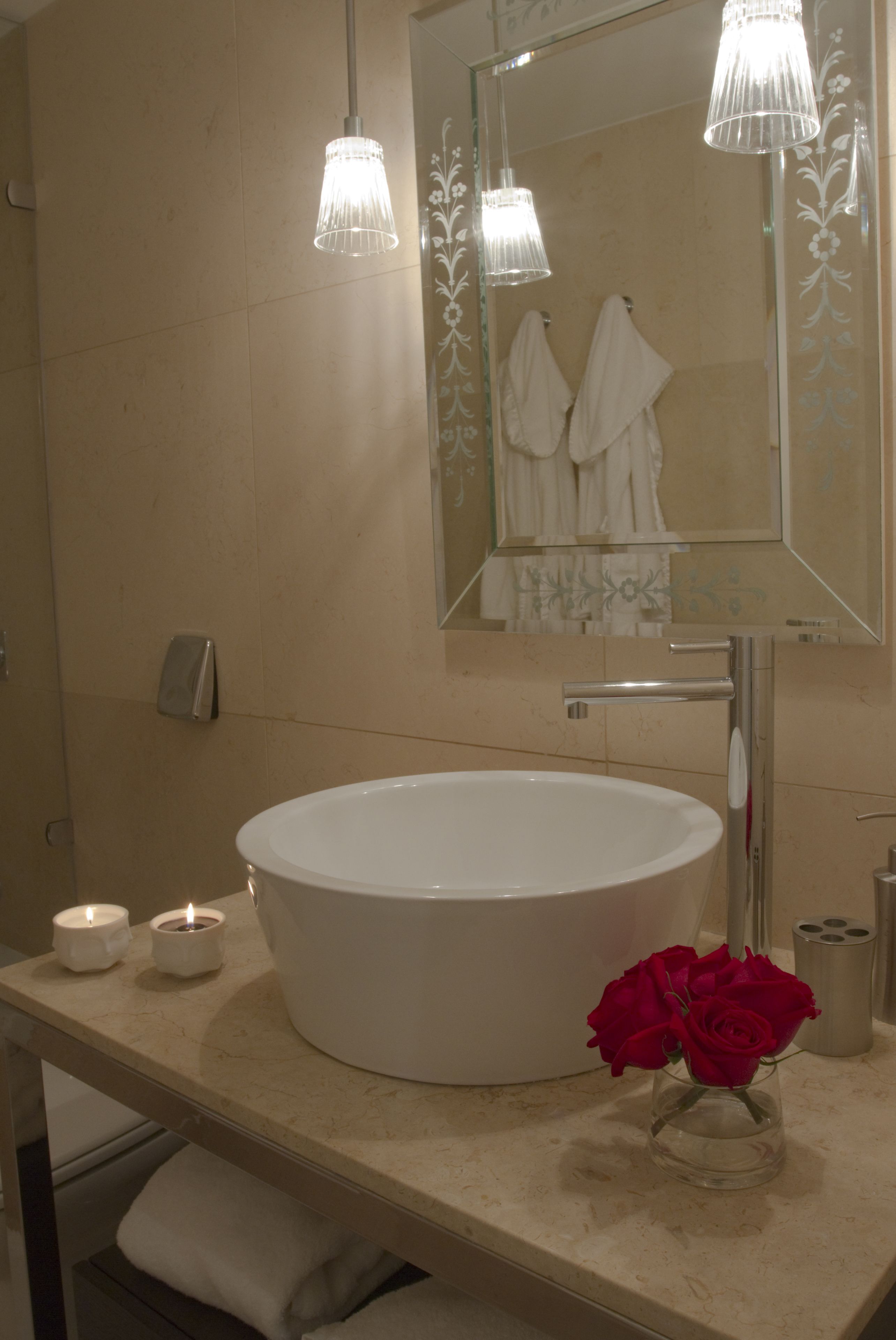 A bathroom with a large white sink, two lit candles, a glass vase with red roses, and a mirror with a floral design.