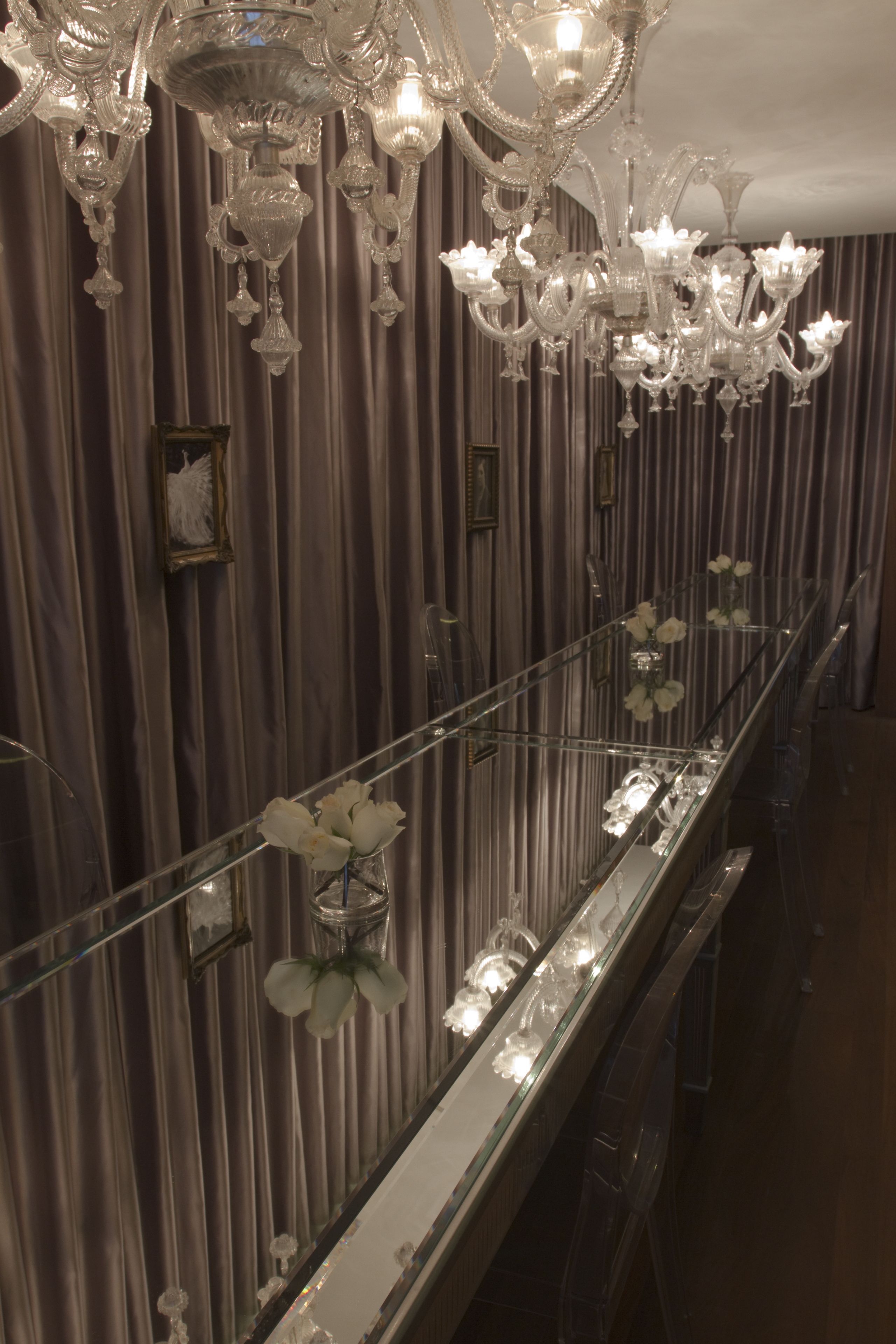 A long, reflective mirror with a glass railing, a chandelier, and a vase of white flowers.