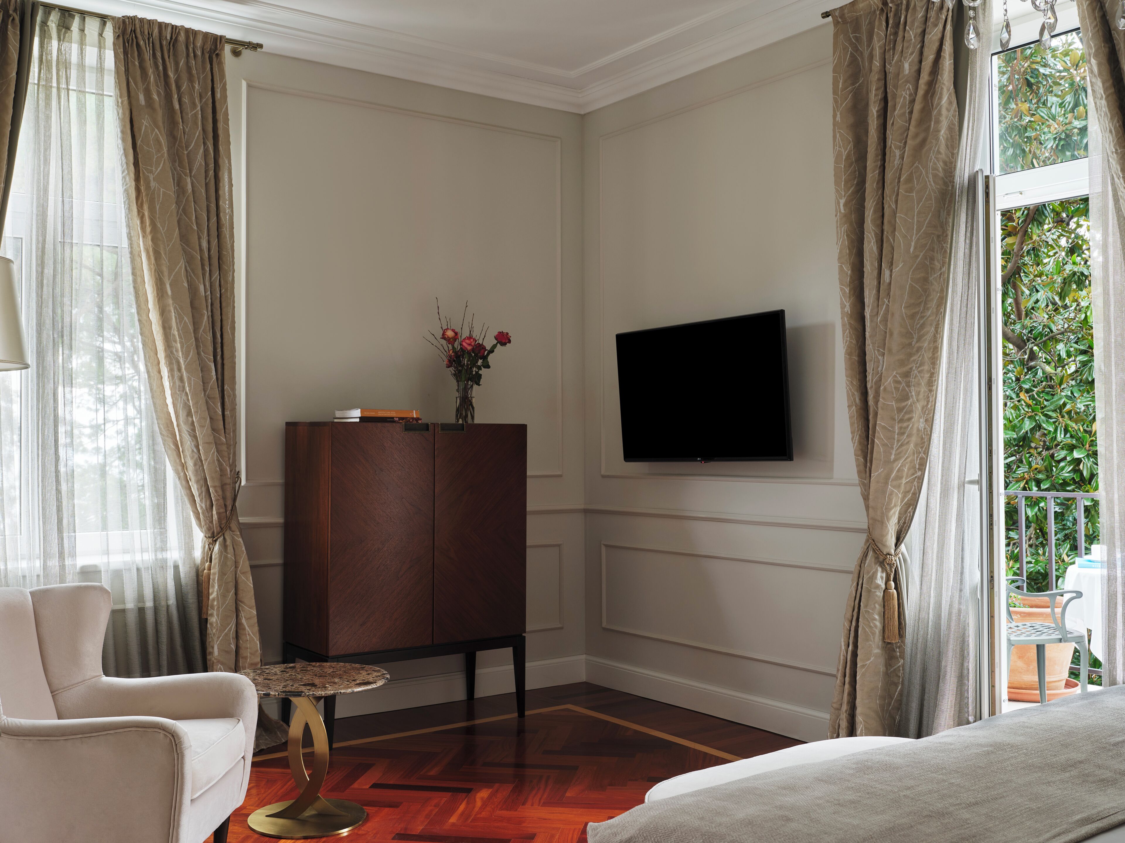 Une chambre d’hôtel avec un meuble en bois, un téléviseur fixé au mur, une table ronde, un canapé et une vue sur la verdure par la fenêtre.