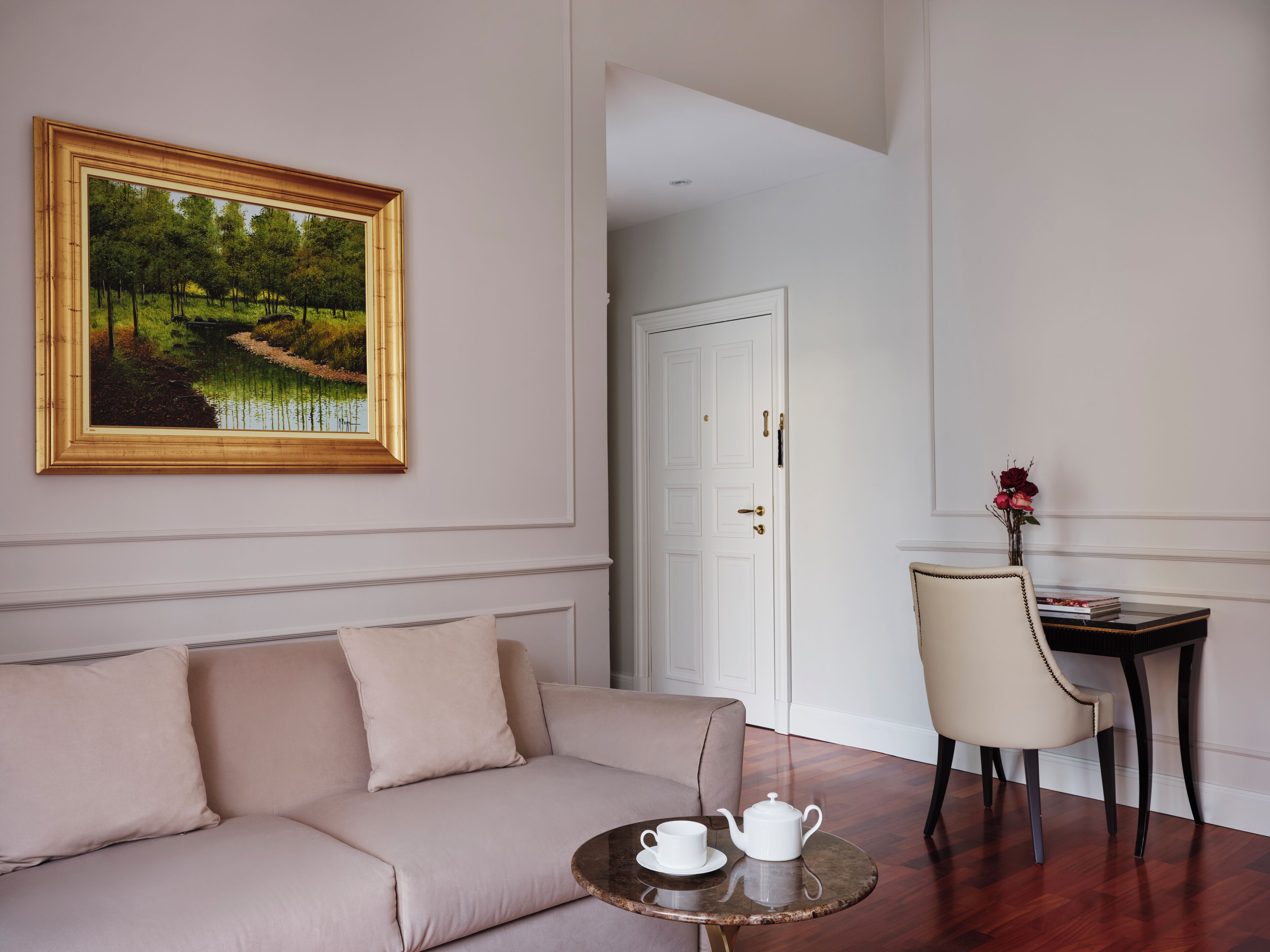 A room with a sofa, a small table with a teapot and cup, a chair, a door, and a framed painting on the wall.