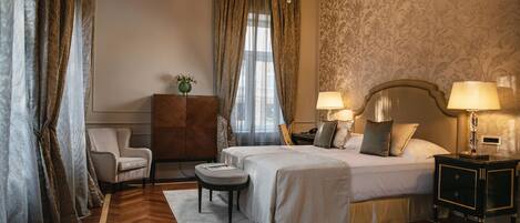 Habitación de hotel con cama, dos lámparas de noche, mesita de noche, sillón, pequeña mesa y candelabro.