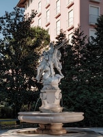A classical stone fountain with a sculpture of figures, surrounded by trees and a pink building in the background.