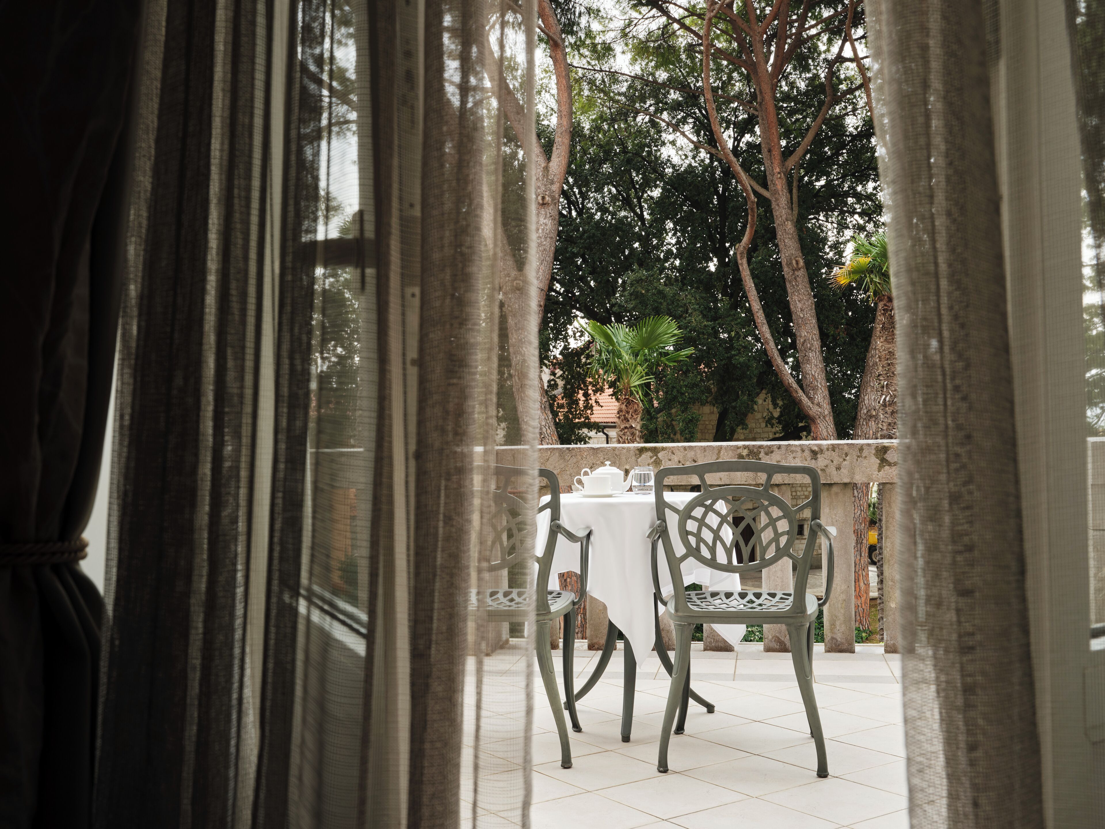 A view through a window with curtains, featuring a table and chairs outside, surrounded by trees.