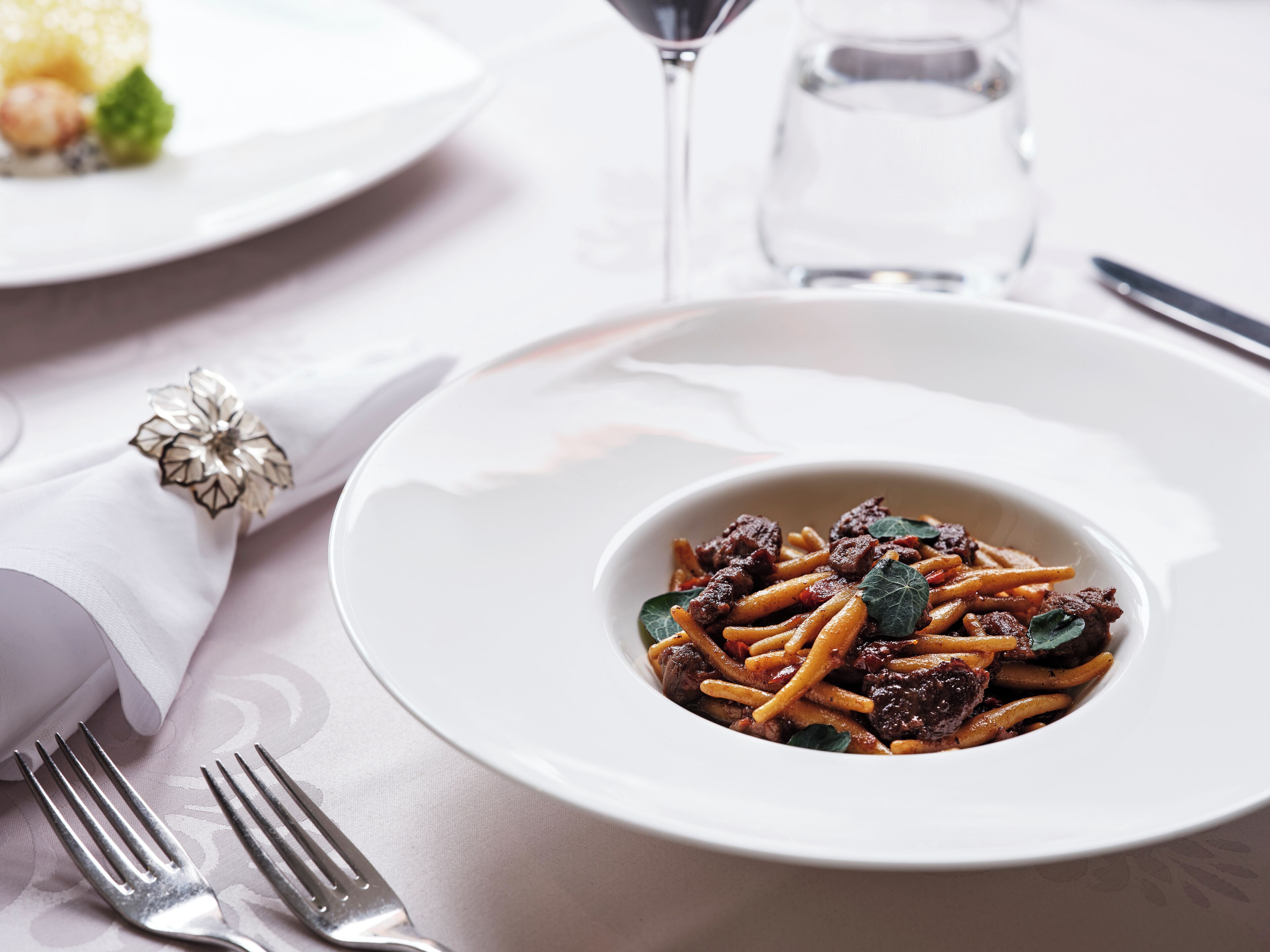 A white plate with pasta and meat, a fork, a glass of water, and a napkin with a silver brooch.
