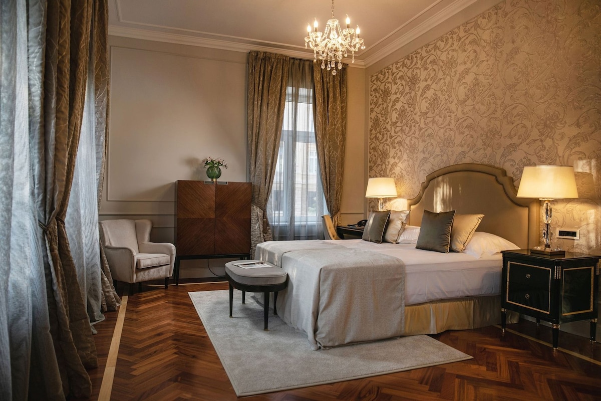 Habitación de hotel con una cama, dos mesitas de noche con lámparas, una silla, una pequeña mesa, un armario y una araña.