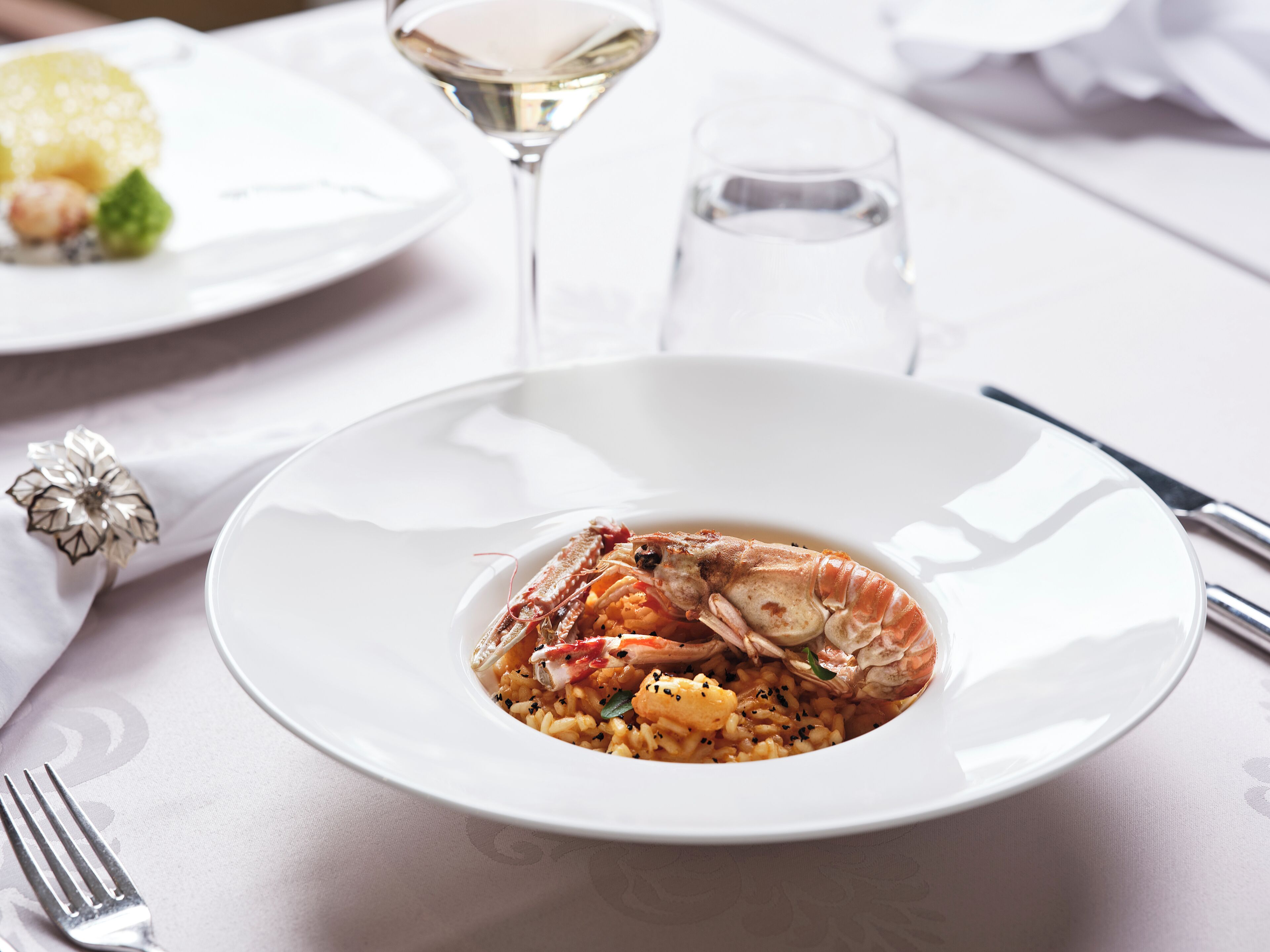 Een bord met zeevruchtenrisotto, met kreeftstaart, vergezeld van een glas witte wijn en een glas water op een tafel.