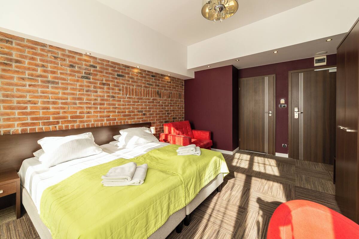 Hotellihuone, jossa on tiiliseinä, vihreillä tekstiileillä varustettu sänky, punaiset tuolit ja puinen vaatekaappi.