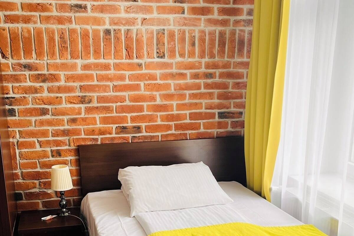 Hotellihuone, jossa on tiiliseinä, valkoisilla lakanoiden ja keltaisella päiväpeitteellä varustettu sänky, yöpöytä, jossa on lamppu, ja ikkuna, jossa on keltaiset verhot.