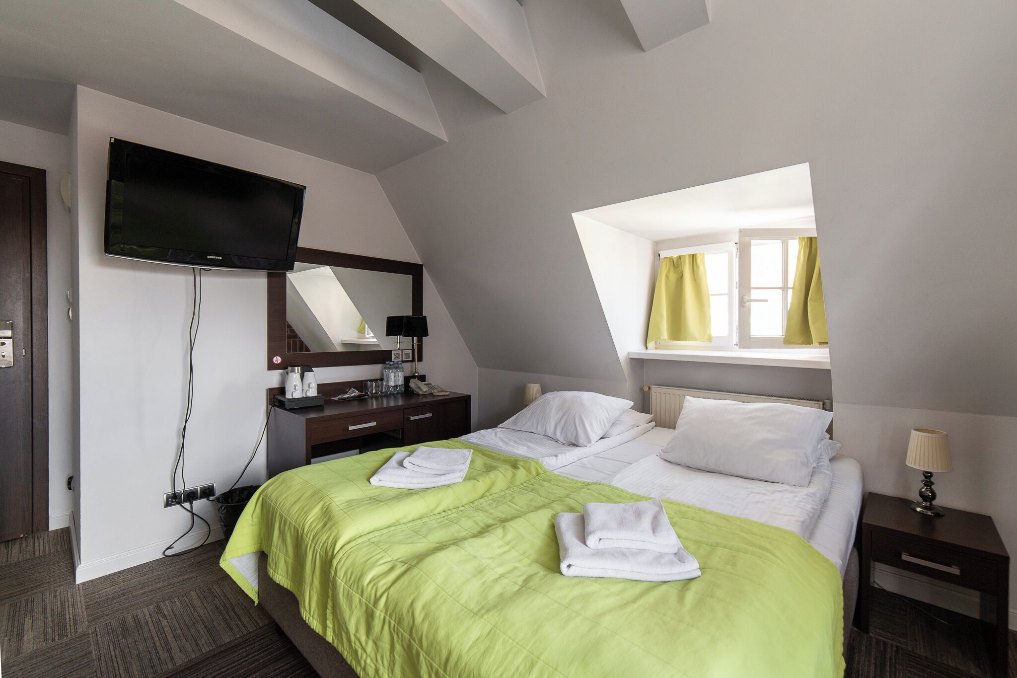 Habitación de hotel con cama, televisor montado en la pared, escritorio con lámpara y ventana con cortinas.