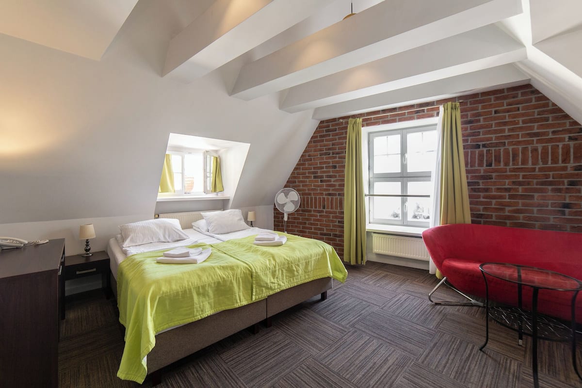 Hotellihuone, jossa on sänky, punainen tuoli, työpöytä, ikkuna verhoilla ja tiiliseinä.