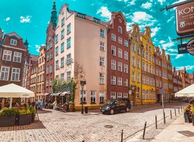 A cobblestone street lined with colorful buildings, a black car parked on the side, and outdoor seating areas with umbrellas.