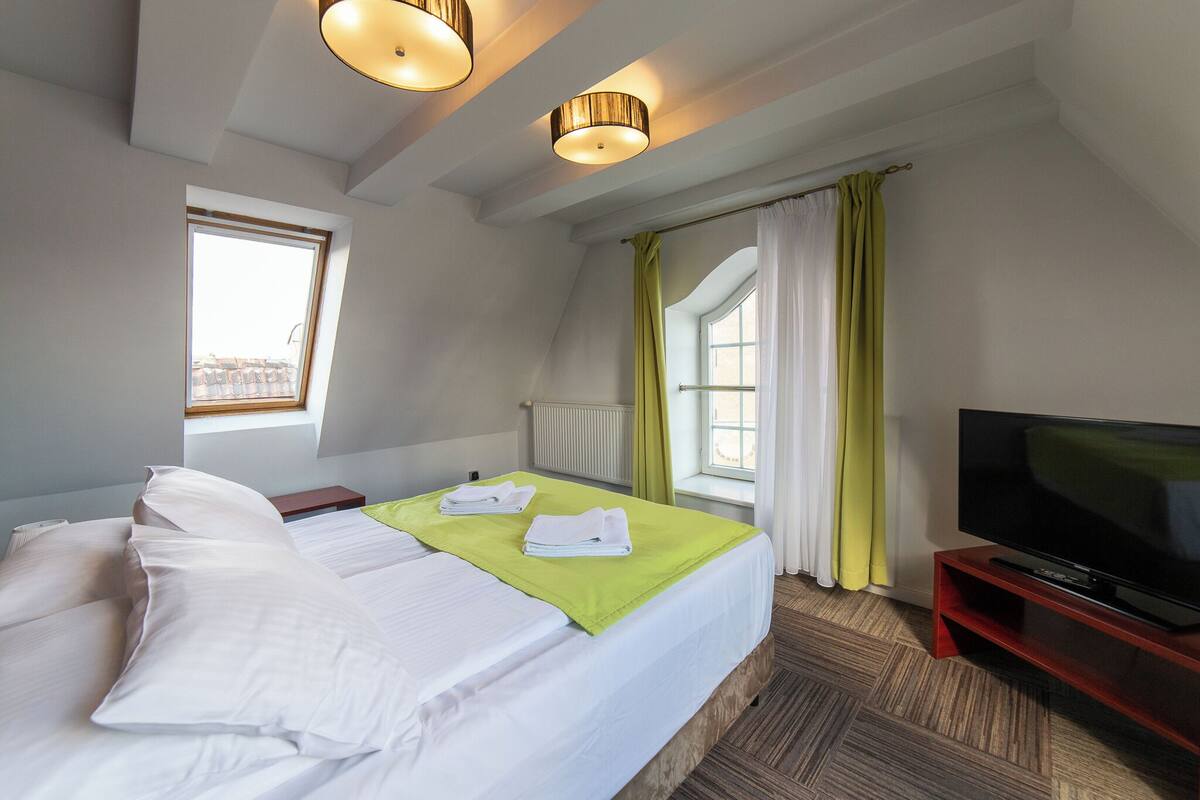 Hotellihuone, jossa on sänky, televisio, ikkuna verhoilla ja kattovalaisin.