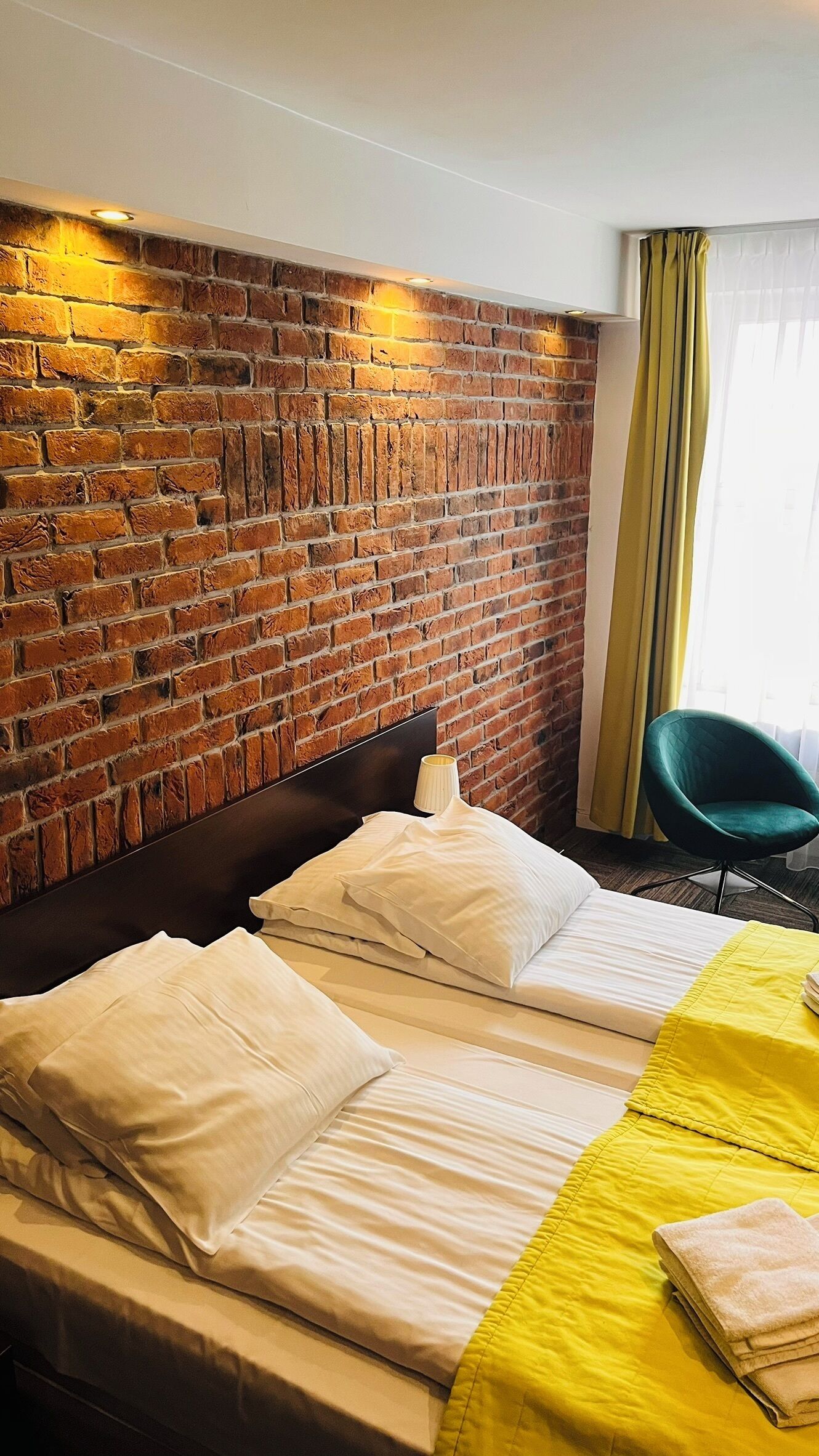 A hotel room with a brick wall, two beds with white linens, a green chair, and a yellow bedspread.