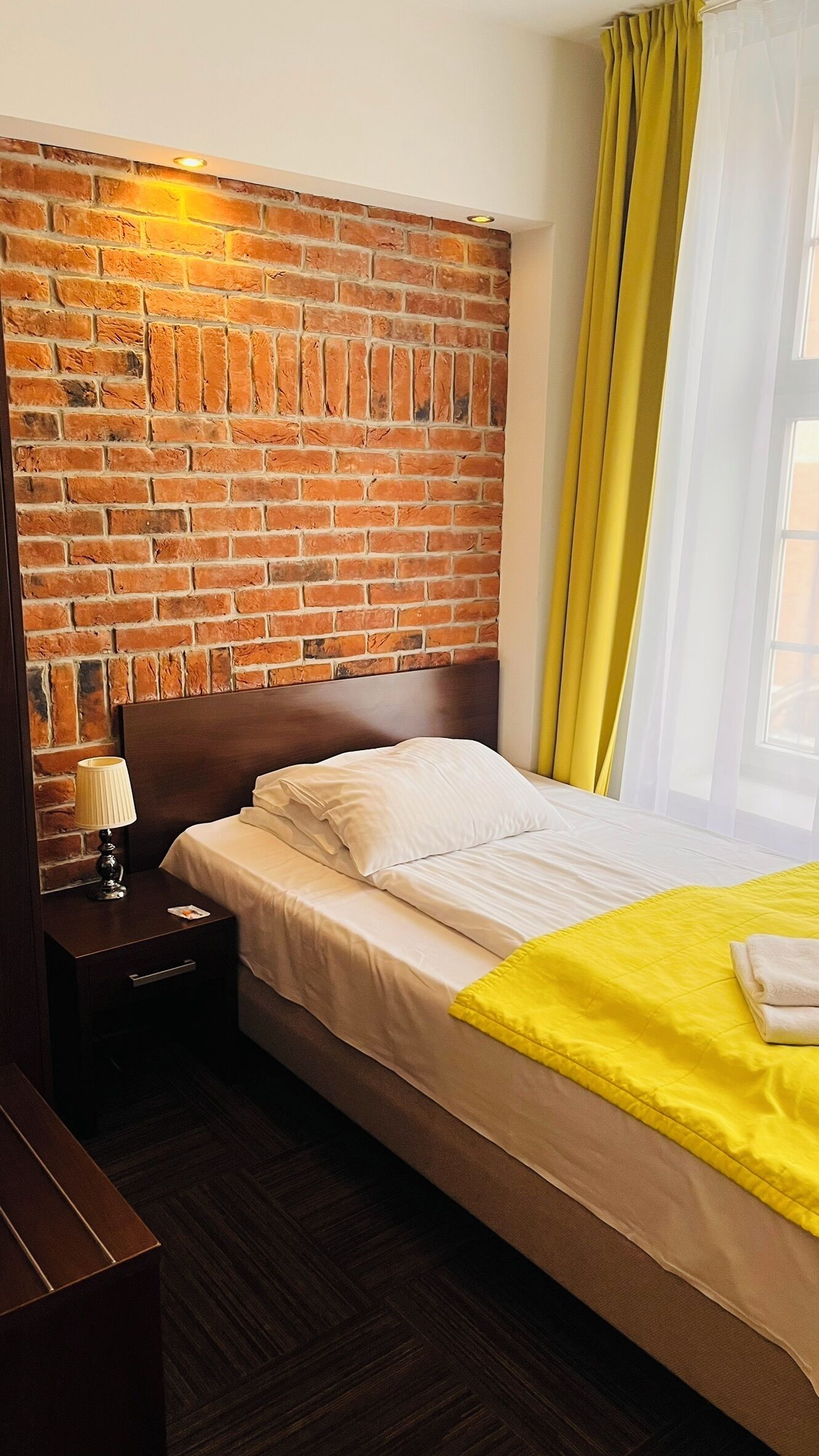 Ein Hotelzimmer mit Backsteinwand, einem hölzernen Kopfteil, einem Bett mit weißer Bettwäsche, einem Nachttisch mit Lampe und einer gelben Decke.