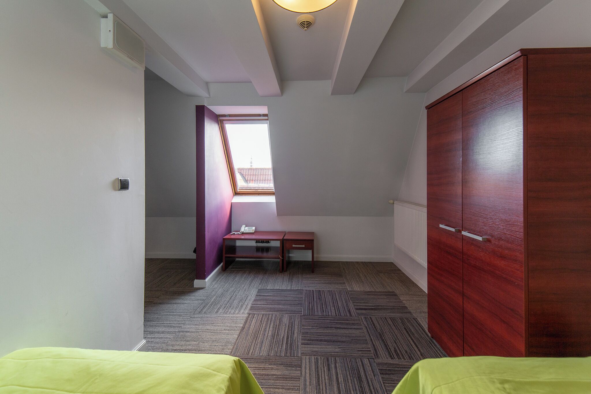 A room with a wooden wardrobe, a desk with a chair, a bed with a green bedspread, and a window with a view of a building.