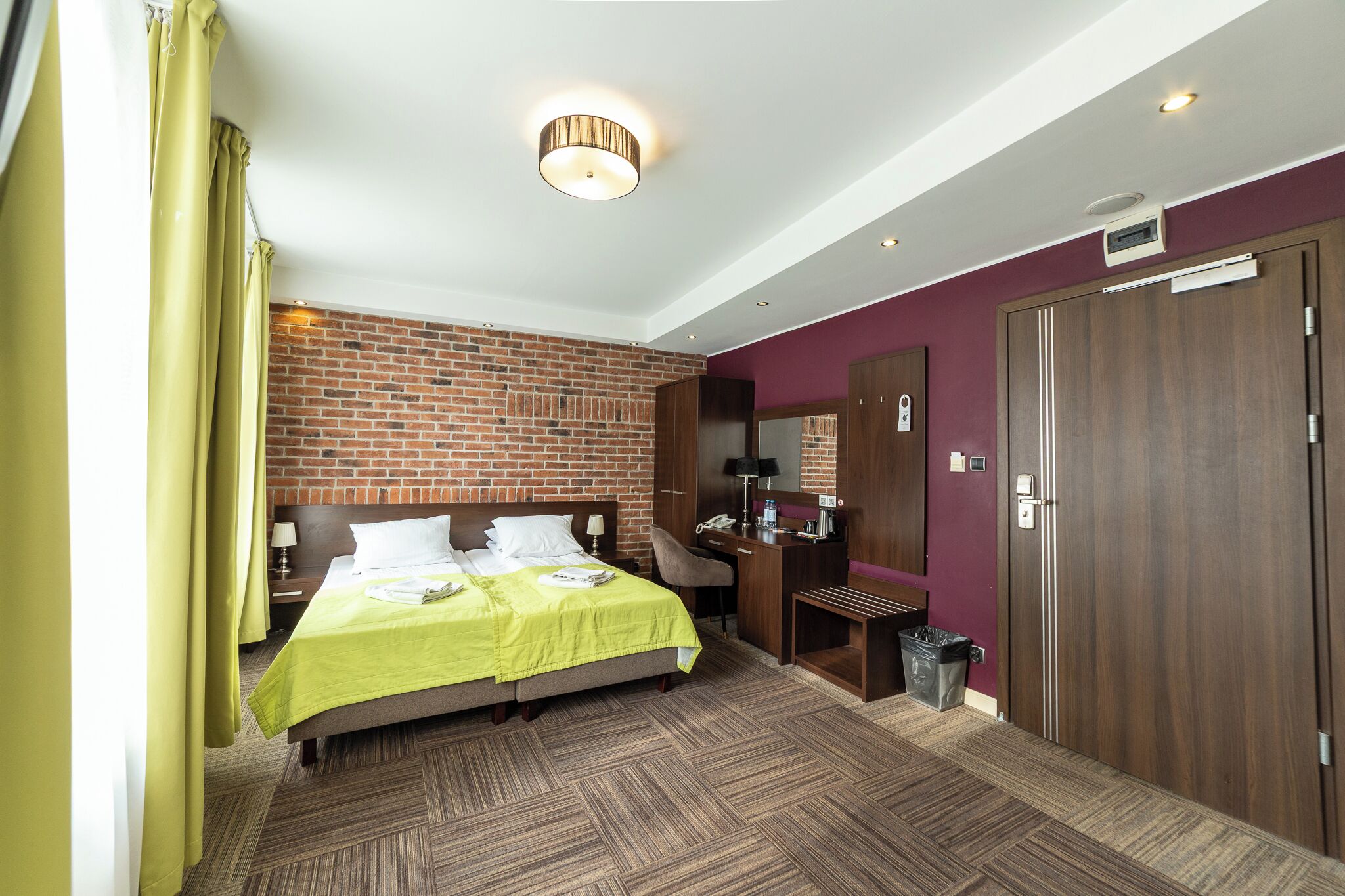 Ein Hotelzimmer mit Backsteinwand, einem Bett mit grüner Bettdecke, einem Schreibtisch mit Computer, einem Stuhl und einem Kleiderschrank.