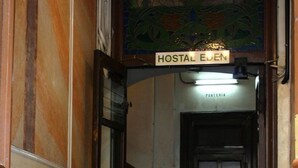 Una mujer sentada en un banco en un pasillo con piso a cuadros, una puerta con la leyenda “HOSTAL EIJEN” y un ventanal de vitral arriba.
