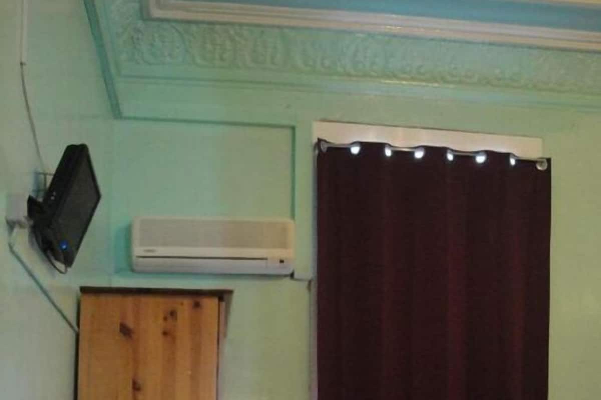 A room with a wooden desk, a bed with a red bedspread, a white air conditioner, and a dark curtain.