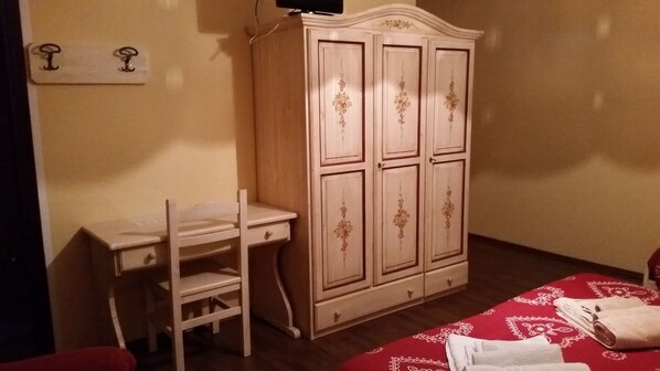 A bedroom with a wooden wardrobe, a desk, and a bed with a red patterned bedspread.